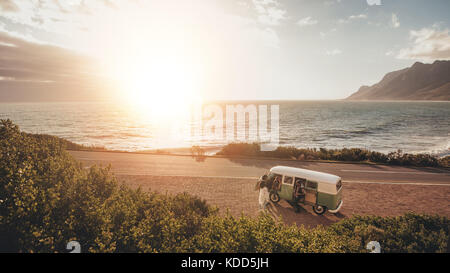 Amici su roadtrip viaggiando da un furgone. Angolo di alta vista del gruppo di persone con il vecchio minivan in piedi lungo la strada costiera durante il tramonto. Foto Stock