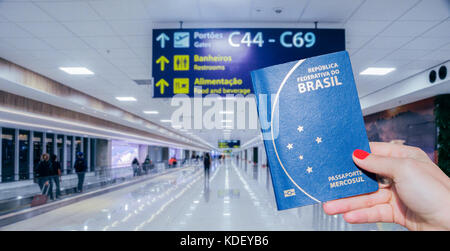 Mano che tiene un passaporto brasiliano e moderno aeroporto terminale e i passeggeri in background Foto Stock