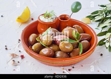 Le olive in un tradizionale ceramica marrone ciotola, con salsa tzatziki, limone, aglio, filiale di olive e di olio d'oliva su un bianco sfondo astratto Foto Stock