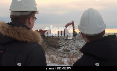 Vista posteriore degli architetti che ispezionano il cantiere. Due ingegneri in elmetti su escavatore da cantiere sullo sfondo. Ingegneri che esaminano i documenti nel cantiere contro l'escavatore. Foto Stock