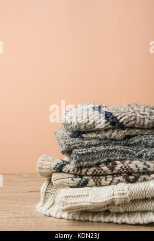 Pila di manufatti a maglia caldo calze sciarpe guanti maglioni di lana grezza e filo marrone grigio beige. tavolo in legno peach colore sfondo parete. eco fashio Foto Stock