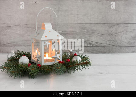 Bianco Natale lanterna con candela accesa su abete ramoscelli decorata con albero di natale baubles su grigio Sfondo legno. Foto Stock