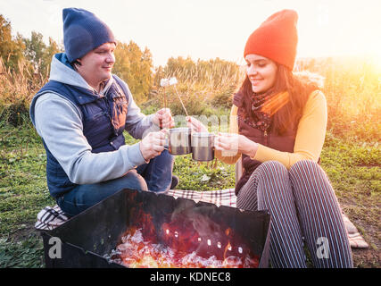 Felice Coppia sorridente e roast marshmallows sul fuoco sulla natura. allegro l uomo e la donna sono in appoggio in corrispondenza del fuoco Foto Stock