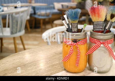 Pennelli in vasi su un tavolo di legno con sfocati i tavoli e le sedie in background Foto Stock