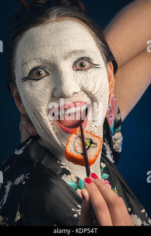 Divertimento ragazza asiatica in abito bianco con incrinato il make-up e zanne su sfondo blu mangiare sushi Foto Stock