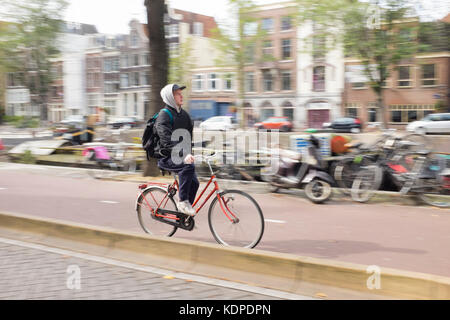 Un giovane casualmente vestito uomo cicli attraverso la zona centrale di Amsterdam Foto Stock