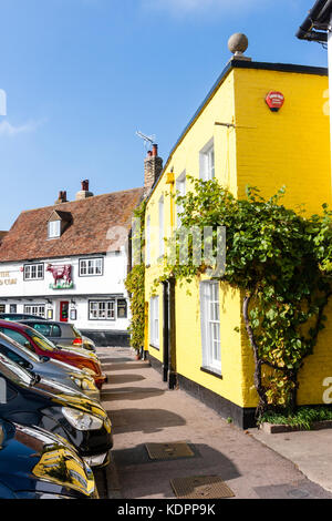 Inghilterra, sandwich. Vista lungo fossato suola marciapiede con auto parcheggiate su un lato e luminosa casa gialla su altri. "Medievale Red Cow' pub in background. Foto Stock