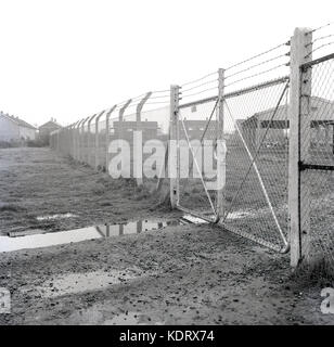 Anni sessanta, storico, Inghilterra, Regno Unito, maglia di catena di recinzione con filo spinato sulla parte superiore e bloccato i cancelli di proteggere e bloccare qualsiasi ingresso illegale di un vecchio industriale cantiere di storage sul bordo di una grande abitazione rurale station wagon. Foto Stock