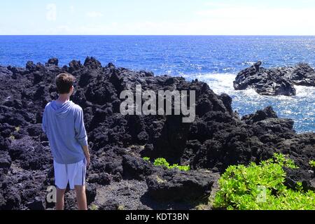 Giovane uomo guarda onde si infrangono in rocce laviche a Waianapanapa State Park, fuori l'Autostrada Hana, Maui, Hawaii Foto Stock