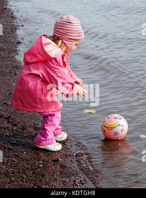 Una bambina di 3 anni nel tentativo di raggiungere la sua palla che ha rotolato nel mare Foto Stock
