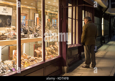 Sera d'inverno nel centro di York, uomo è in piedi guardando orologi illuminato & sul display in smart gioiellerie finestra - Stonegate, Inghilterra, Regno Unito. Foto Stock