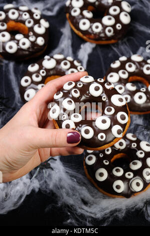 Mano trattiene una ciambella con glassa di cioccolato decorate googly occhi su halloween Foto Stock