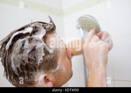 Un giovane uomo con una testa di sapone è in piedi nel bagno e guarda la doccia con perplessità. Foto Stock