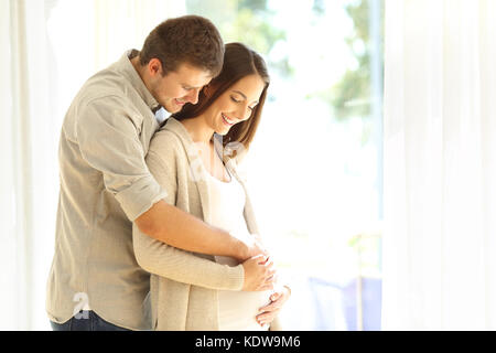 Orgogliosi i nuovi genitori guardando il ventre della donna incinta in piedi accanto a una finestra a casa Foto Stock