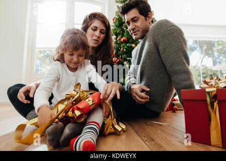 Il padre e la madre guarda il loro bambino aprire il suo regalo di Natale. Albero di Natale decorato e confezioni regalo in background. Foto Stock