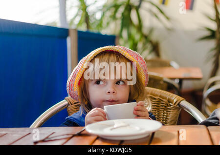Little White Caucasian ragazza bionda indossando Rosa Estate Hat seduti al tavolo a bere il latte caldo dal bianco coppa in ceramica come un adulto in Alfresco Di Foto Stock