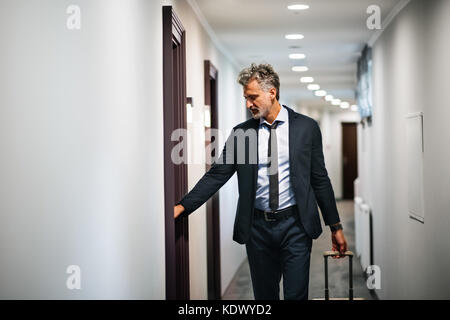 Imprenditore maturo con i bagagli in un hotel corridoio, aprendo la porta della camera uomo bello tirando la valigia. Foto Stock