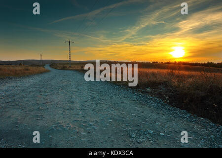 Vista di una strada rurale al tramonto, bellissimi colori arancione all'orizzonte Foto Stock