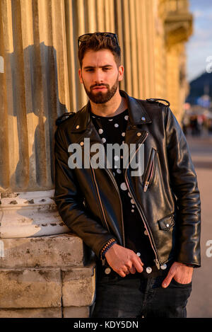 Un bel giovane hipster appoggiato alla colonna di una casa su una strada soleggiata e rilassante Foto Stock