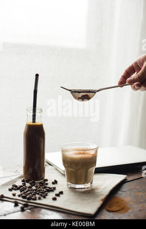 Barattolo di caffè drink con tubo e bicchiere di cappuccino con cacao in polvere sul tovagliolo con chicchi di caffè Foto Stock