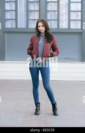 Giovane bella donna elegante in un pilota di borgogna giacca e jeans blu, street style, Primavera e autunno trend, guardare, capelli al vento Foto Stock