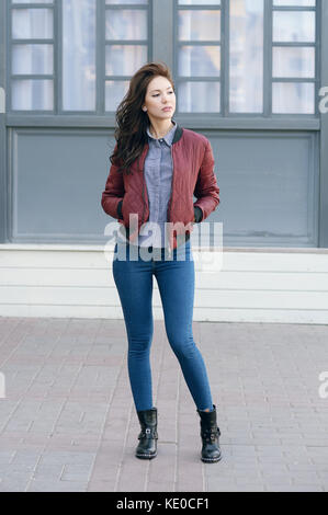 Giovane bella donna elegante in un pilota di borgogna giacca e jeans blu, street style, Primavera e autunno trend, guardare, capelli al vento Foto Stock