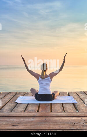 Vista posteriore del giovane donna sat su asciugamano a praticare yoga dal mare al tramonto Foto Stock