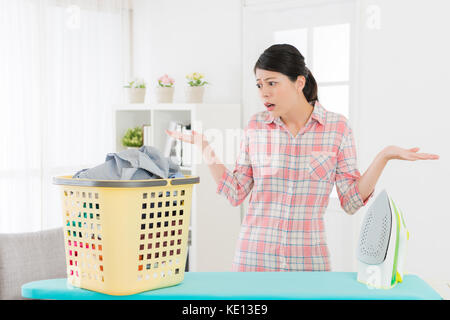 Piuttosto elegante studentessa guardando abbigliamento disordinato e mostrando ammutolito emotivo con gesto quando ha sempre bisogno di aiutare la madre a stirare cl Foto Stock