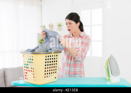 Bella donna dolce tenuta camicia grigia guardando area sporca sensazione sconvolto con cesto per la biancheria e asse da stiro nel salotto di casa. Foto Stock