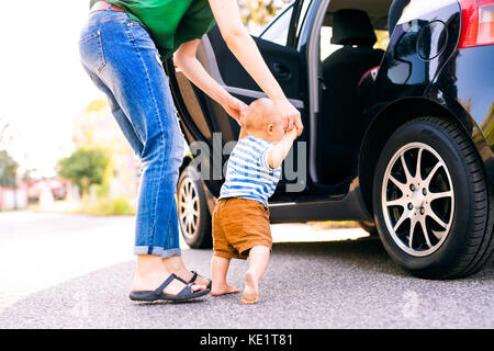 Irriconoscibile giovane madre aiutando il suo piccolo bimbo a piedi. Primi passi, nei pressi di un'auto. Foto Stock