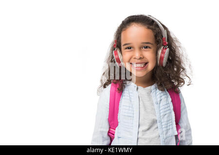 Adorable African American schoolgirl ascoltando musica in cuffia e sorridente in telecamera isolato su bianco Foto Stock