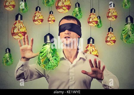 Gli occhi bendati giovane uomo a piedi attraverso le lampadine a forma di cibo spazzatura e verdura verde isolato sul muro grigio Sfondo. dieta scelta nutriti di destra Foto Stock