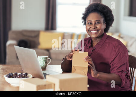 Sorridente giovane donna africana seduti ad un tavolo a casa con un computer portatile e gli indirizzi di scrittura a pacchetti per la sua home base business online Foto Stock