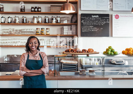 Ritratto di un sorridente giovane africano imprenditore femmina in piedi con le braccia incrociate al bancone del suo bistro Foto Stock