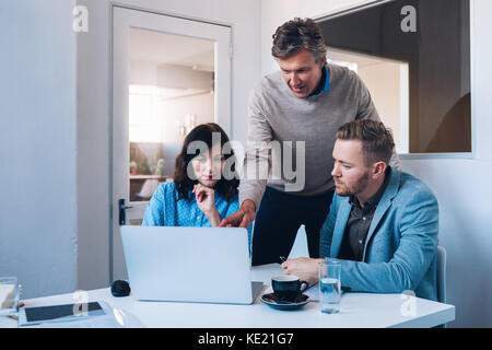 Manager che offre consulenza in piedi dietro due giovani colleghi seduti ad un tavolo insieme al lavoro su un notebook in un luminoso ufficio moderno Foto Stock