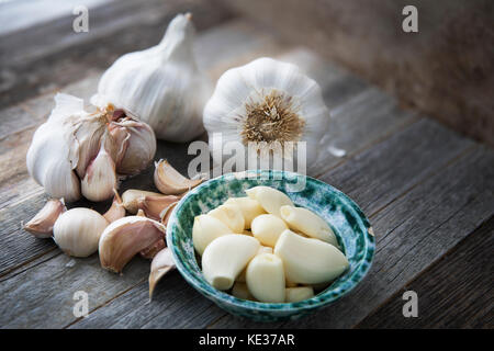 Ciotola di spicchi di aglio sgusciati in ciotola con più i chiodi di garofano e teste d'aglio su una superficie di legno Foto Stock