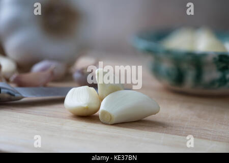 Spicchi di aglio sgusciati sul tagliere di legno con coltello e ciotola in background. Foto Stock