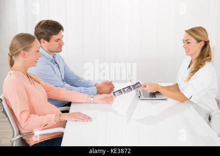 Coppia giovane guardando a ultrasuoni report di scansione mostrato dal medico con laptop sulla scrivania Foto Stock