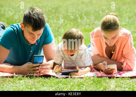 Genitori felici che stabilisce nel Parco guardando al suo figlio utilizza lo smartphone Foto Stock