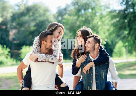 Due coppie amorevole piggyback avendo divertimento all'aperto. le ragazze seduta sul retro dei ragazzi sorridenti. giovani uomini e donne a giocare nel parco e guardando ogni altri Foto Stock