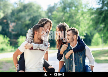 Due coppie amorevole piggyback avendo divertimento all'aperto. le ragazze seduta sul retro dei ragazzi sorridenti. giovani uomini e donne a giocare nel parco e guardando ogni altri Foto Stock