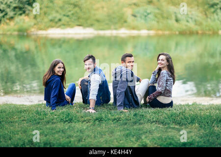 Due coppie seduto sull'erba dal fiume rivolta alla ricerca della fotocamera. quattro giovani seduta dal lago a mezzo giro e guardando indietro. Gli studenti rilassarsi in romant Foto Stock