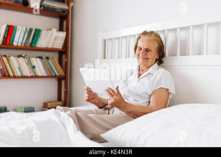 Donna anziana comodamente seduto sul letto e usando la sua tavoletta digitale Foto Stock