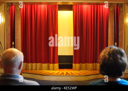 Il velluto tende rosse leggermente aperto mostra un uomo e una donna guarda in un cinema privato costruito nel 1920, inizio stile anni trenta. Illuminata luminosamente. Foto Stock