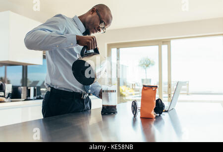 Uomo che versa acqua calda nella macchina per il caffè. uomo d'affari che prepara il caffè al tavolo della colazione con un computer portatile accanto. Foto Stock