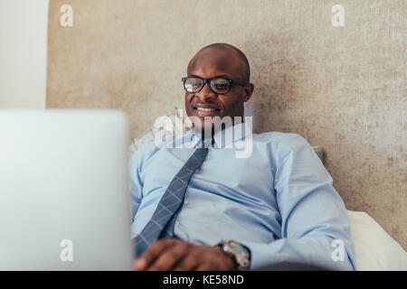Uomo in abiti formali lavorando sul computer portatile mentre giaceva a letto. Uomo sorridente lavorando su laptop rilassante sul letto. Foto Stock