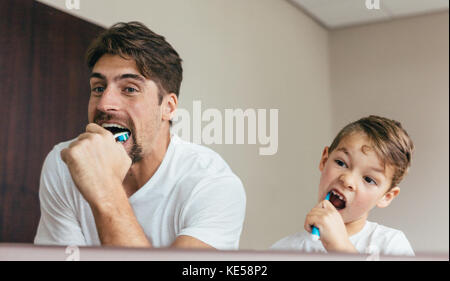 Padre e figlio di spazzolare i denti in bagno. Giovane uomo con suo figlio insieme la spazzolatura dei denti e cercando in mirror. Foto Stock