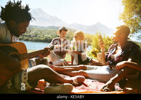 I giovani amici si esibono, suonano la chitarra e si gustano un picnic sul soleggiato lungofiume estivo Foto Stock