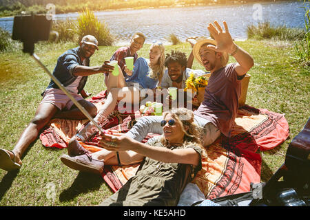 Giocosi giovani amici che prendono selfie con bastone selfie, gustando pic-nic in estate soleggiato lungofiume Foto Stock