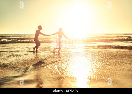 Giovane coppia che tiene le mani, camminando in estate soleggiato tramonto mare spiaggia surf Foto Stock